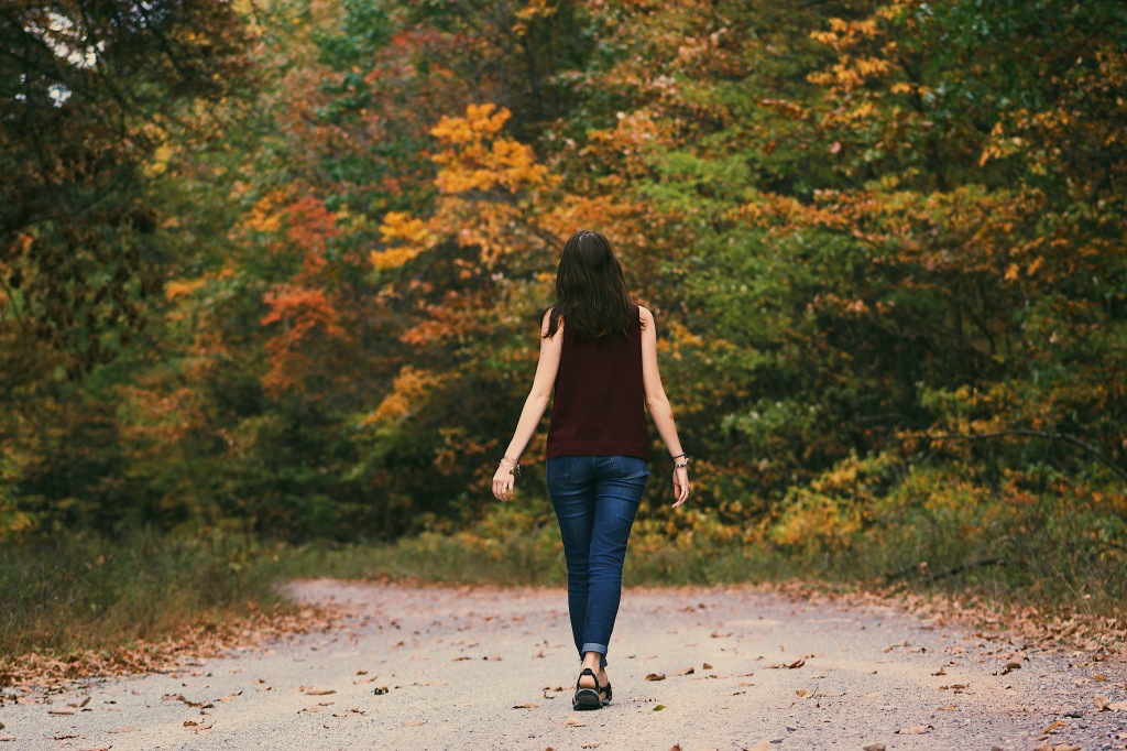 A woman seen from behind, walking on a wide path surrounded by trees in the fall.